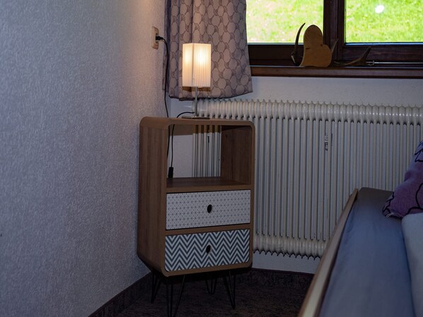 A corner bedroom with a wooden bedside table, a lamp, a radiator, and a window with curtains.