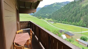 Ein Holzbalkon mit Tisch und Stuhl, der auf ein grünes Tal und bewaldete Hügel blickt.