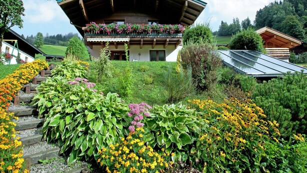 Ein traditionelles Chalet mit geneigtem Dach, einem Balkon voller Blumen und einem Garten mit verschiedenen Pflanzen und Blumen.