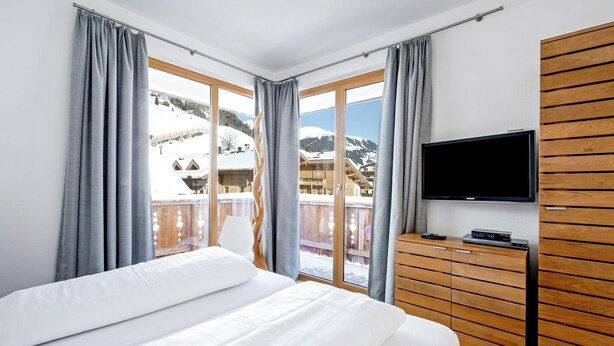 Habitación de hotel con cama, televisión, cómoda de madera y balcón con vistas a montañas nevadas.