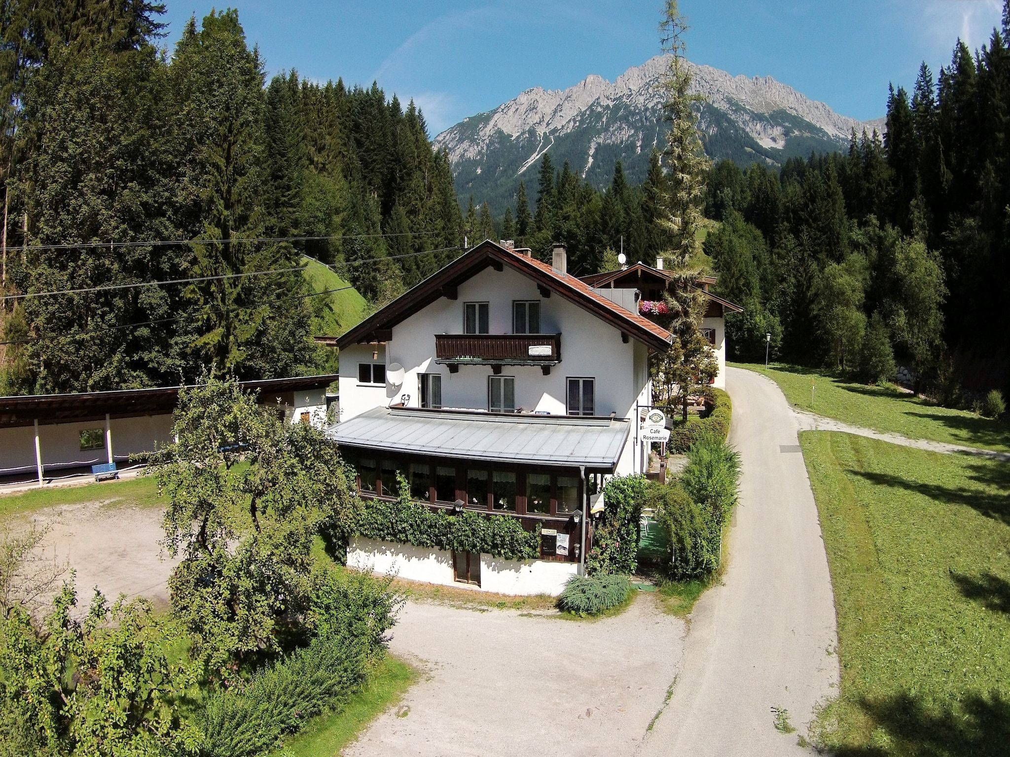 A two-story chalet with a balcony, surrounded by lush greenery and a mountain backdrop.