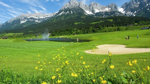 Un parcours de golf avec un étang, une fontaine et des montagnes en arrière-plan.