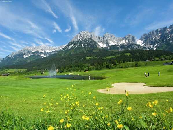 Un campo de golf con un estanque, una fuente y montañas al fondo.