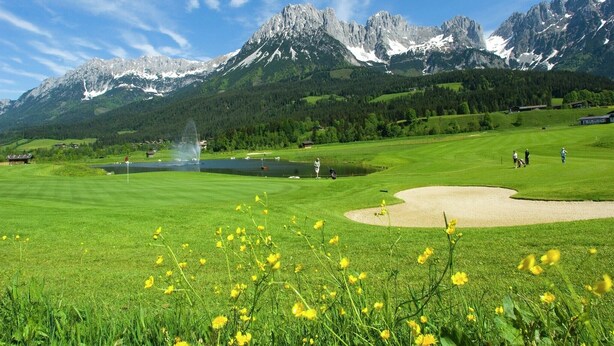 Un campo de golf con un estanque, una fuente y montañas al fondo.
