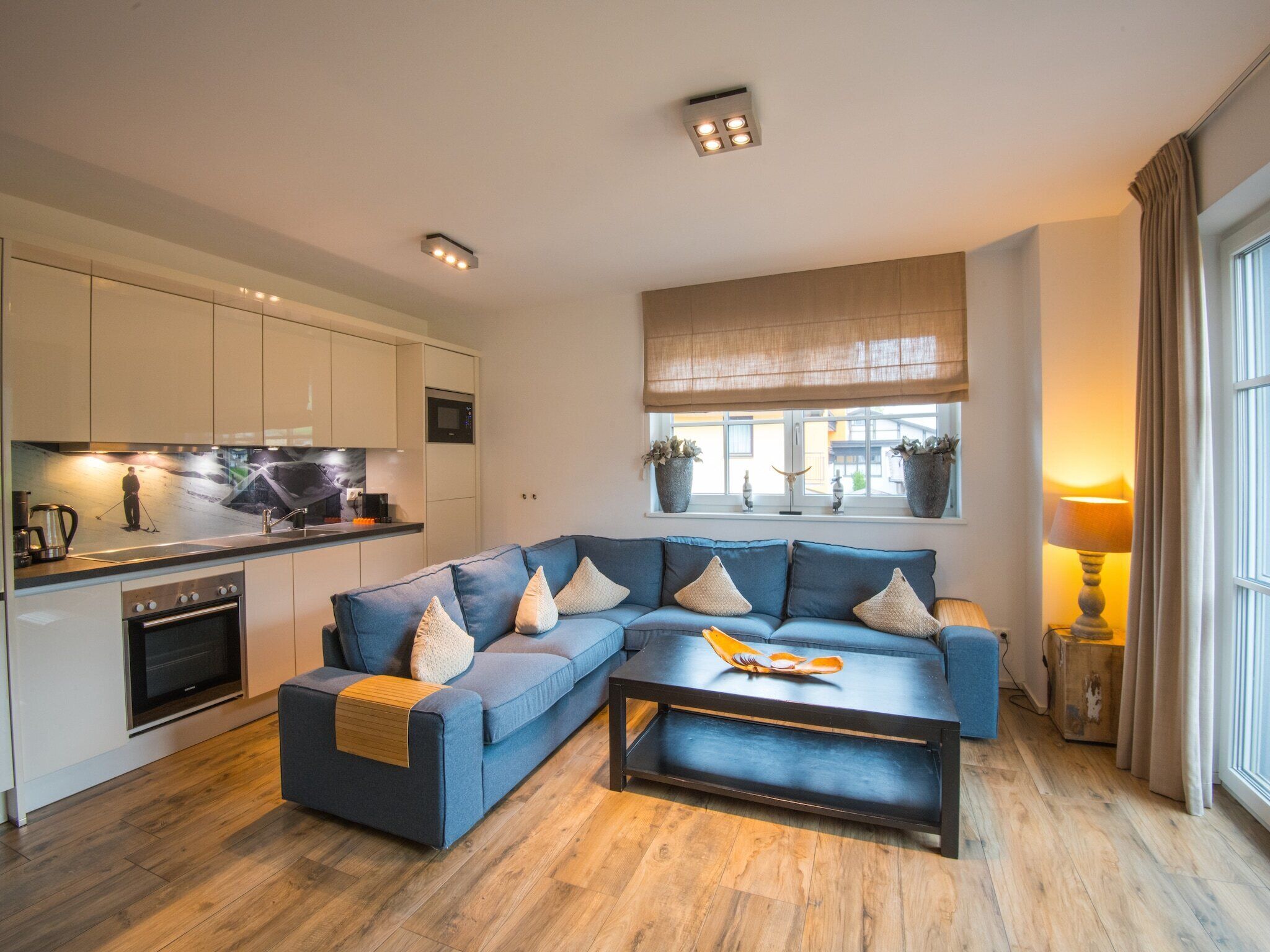 A modern living room with a sectional sofa, a coffee table, and a kitchen area with a stove and cabinets.