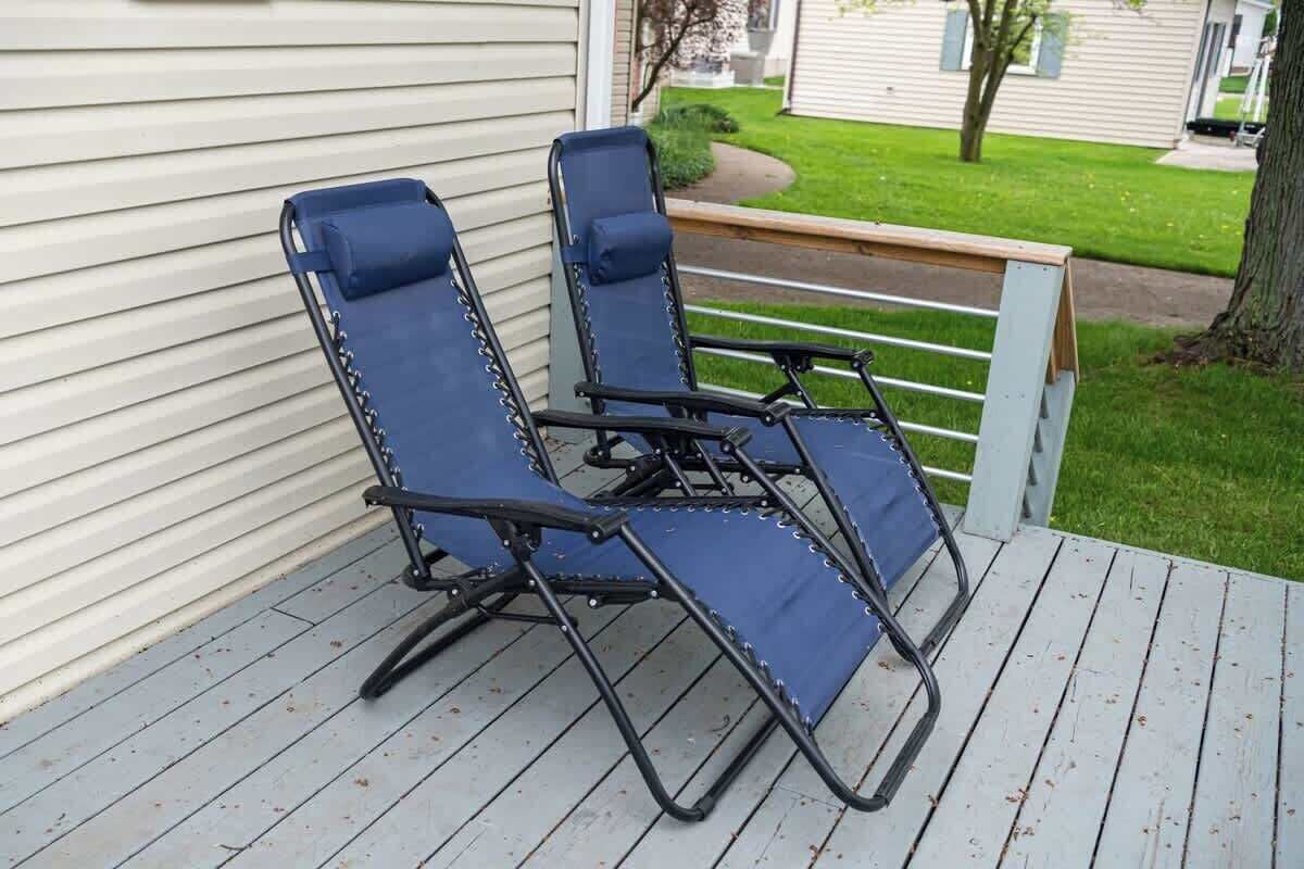 Two blue lounge chairs on a wooden deck with a white railing, a grassy yard, and a house in the background.