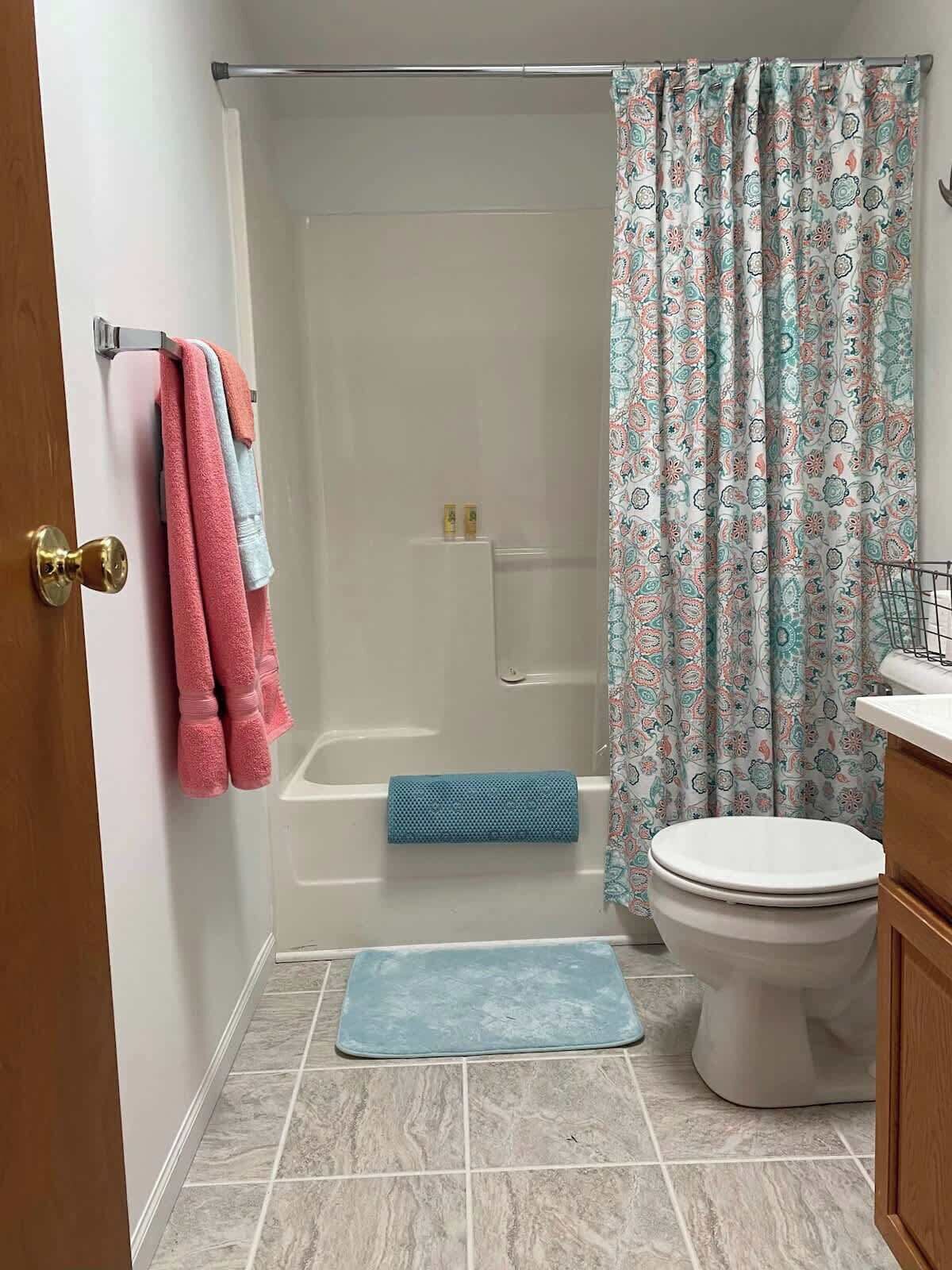 Un baño con ducha, inodoro y lavamanos. Hay toallas colgadas en un perchero y una cortina de ducha con estampado floral.