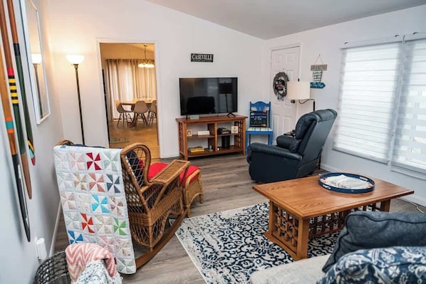 Ein Wohnzimmer mit Fernseher, einem blauen Sessel, einem Holztisch und einem gemusterten Teppich.