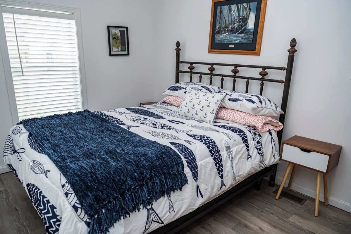 A neatly made bed with a patterned bedspread and pillows, a bedside table with a drawer, and a framed picture on the wall.
