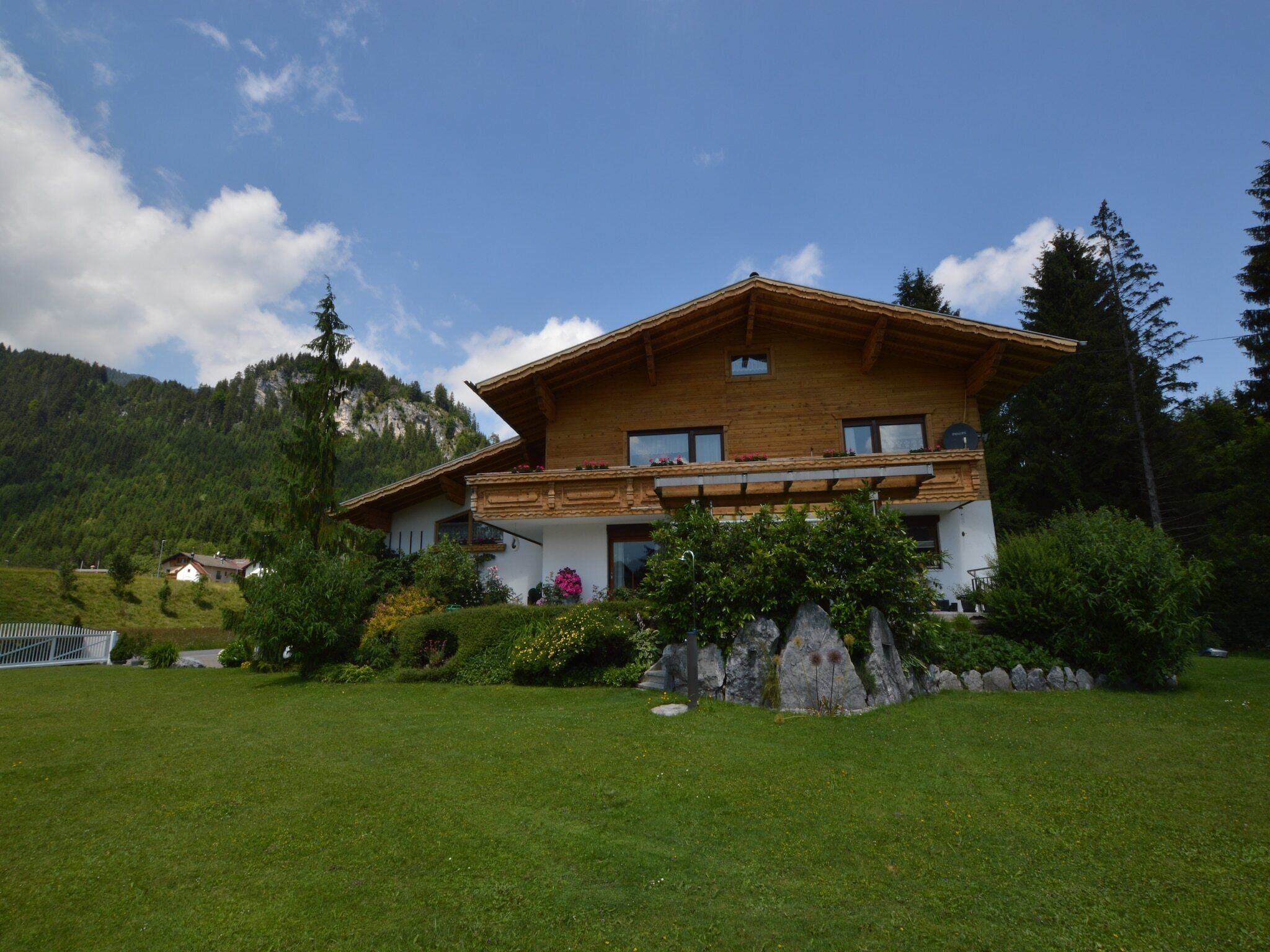 A two-story wooden house with a balcony, surrounded by a well-maintained lawn and a mountain backdrop.