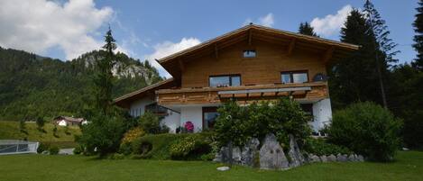 A two-story wooden house with a balcony, surrounded by a well-maintained lawn and a mountain backdrop.