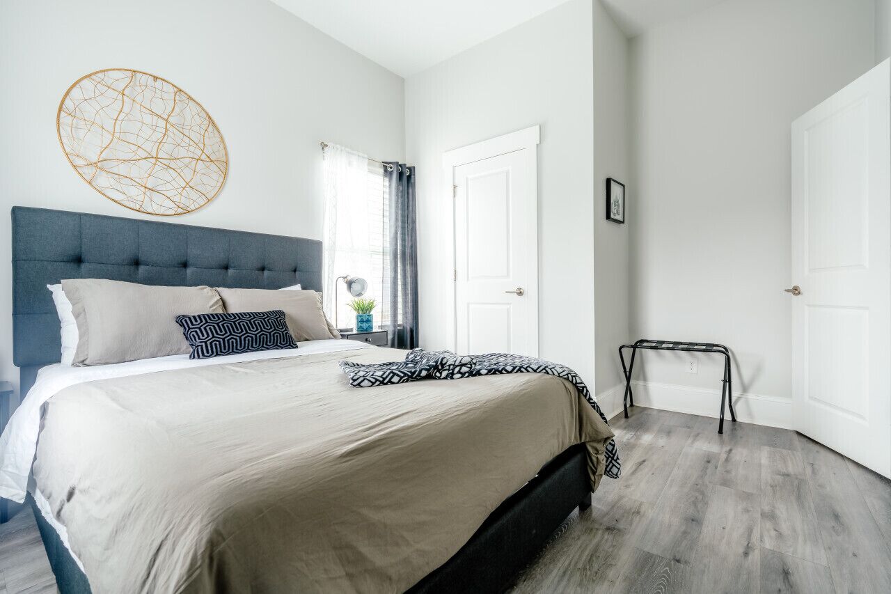 A bedroom with a tufted headboard, a large bed with beige bedding, a geometric wall art piece, a window with curtains, and a door.