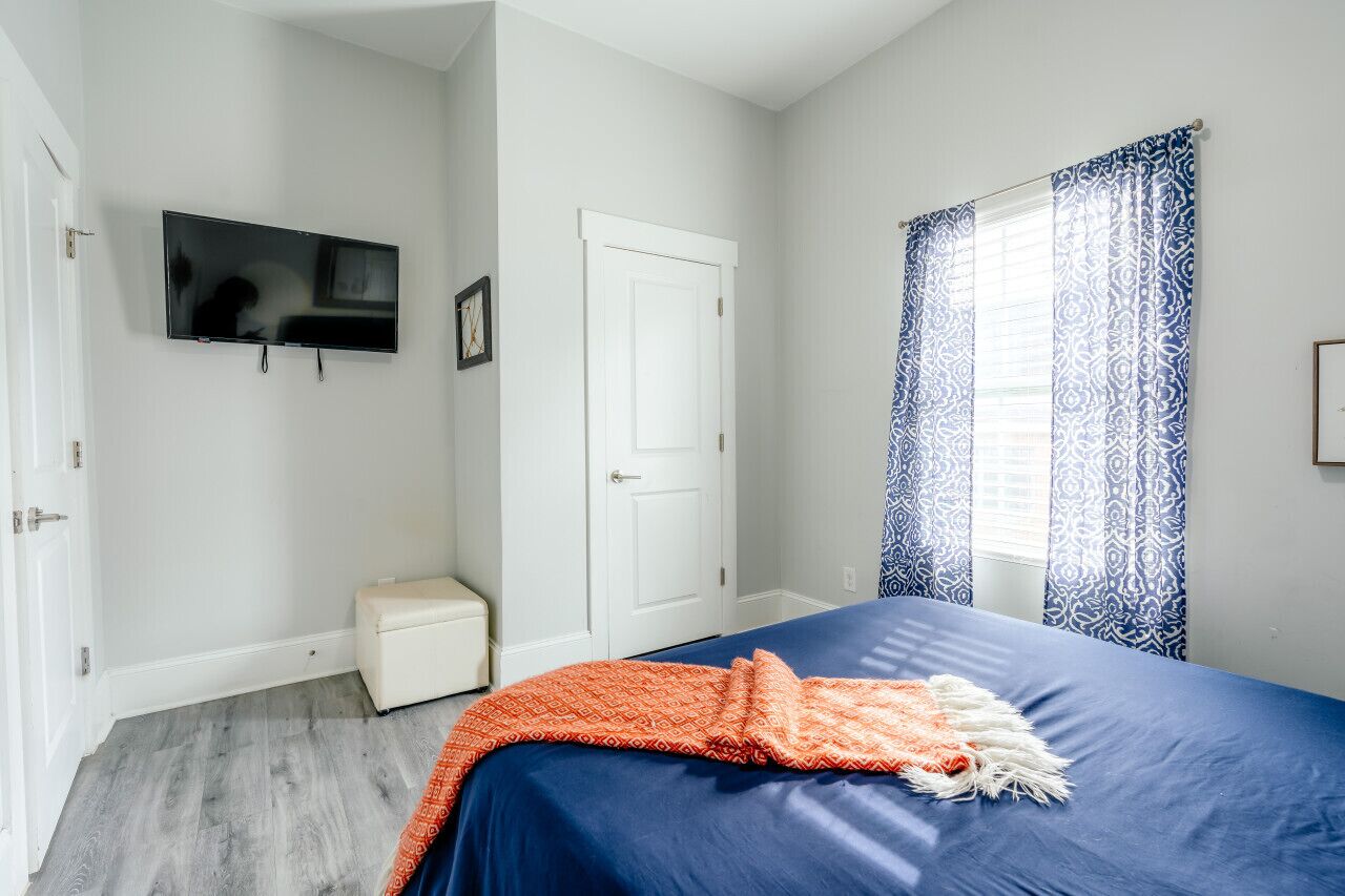 A bedroom with a bed, a television on the wall, a white door, and a window with patterned curtains.