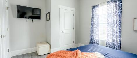 A bedroom with a bed, a television on the wall, a white door, and a window with patterned curtains.