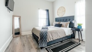 Un dormitorio con cama, televisor montado en la pared, mesita de noche con lámpara y ventana con cortinas.