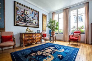 Ein Zimmer mit einem großen abstrakten Gemälde, einer Holzkommode, einem blauen Teppich mit Muster und zwei roten Stühlen.
