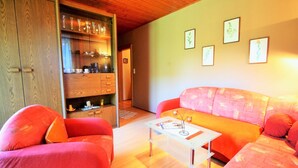 A cozy living room with a wooden ceiling, a red sofa, a glass coffee table, and a wooden cabinet with glassware.