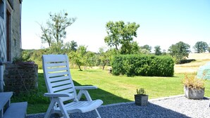 Un'area relax esterna con sedute, un vialetto in ghiaia, un piccolo vaso con una pianta e vista su un paesaggio verde con alberi e siepe.