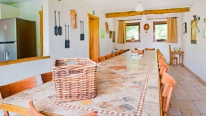 Una larga mesa de comedor con sillas, una cesta tejida y una zona de cocina con refrigerador.
