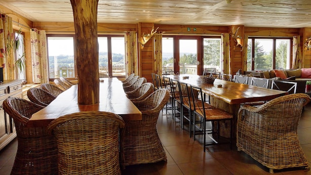 A spacious dining area with a long wooden table and wicker chairs, large windows offering a view of the outdoors, and a rustic ceiling. - Luxurious Holiday Home in Stoumont with Pool & Bar (Stoumont)