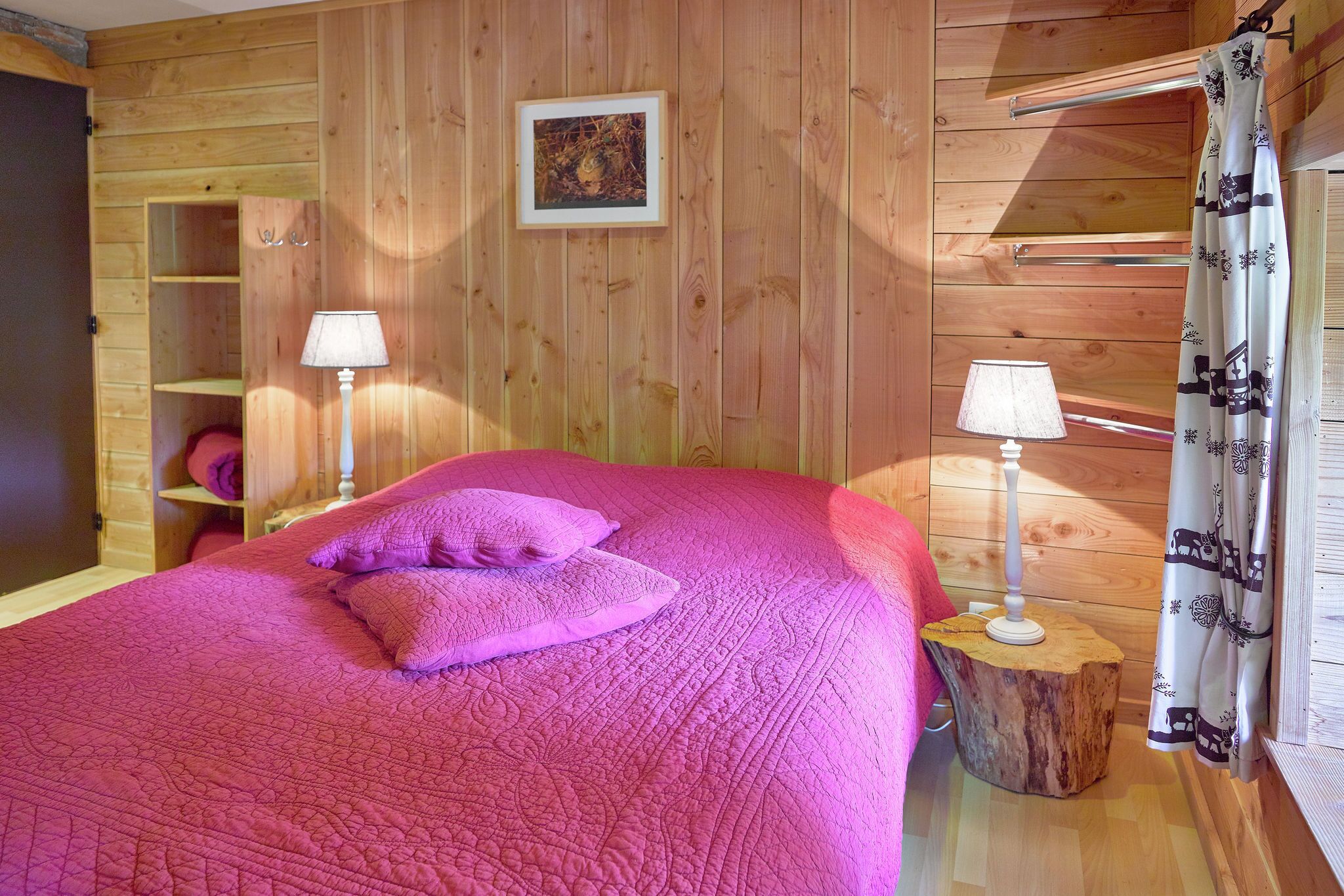Ein Schlafzimmer einer Holzhütte mit einem Bett, zwei Lampen, einem Bild an der Wand und einem Fenster mit Vorhängen.