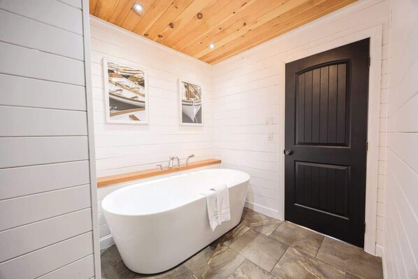A bathroom with a freestanding bathtub, wooden ceiling, and a dark door.