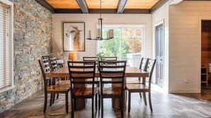 Een eetgedeelte met een houten tafel en stoelen, een stenen muur en uitzicht op bomen door een raam.