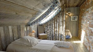 A rustic bedroom with a wooden ceiling, a sloped roof, a bed with white linens, a small bedside table with a lamp, and a brick wall. - Comfortable chalet with indoor pool, hammam and sauna near Stoumont (Stoumont)