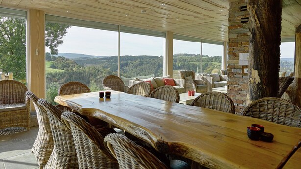 A dining area with a long wooden table, wicker chairs, and a view of a forested landscape through large windows. - Comfortable chalet with indoor pool, hammam and sauna near Stoumont (Stoumont)