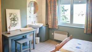 A compact bedroom with a single bed, a bedside table, a small desk with a chair, a window with curtains, a radiator, and a framed floral artwork on the wall. - Spacious, atmospheric upper floor of a farmhouse with garden (Loenhout)