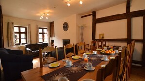 Ein Essbereich mit einem Holztisch, der für das Frühstück gedeckt ist, umgeben von Stühlen. Eine Uhr ist an der Wand zu sehen, und ein Fenster lässt natürliches Licht herein.