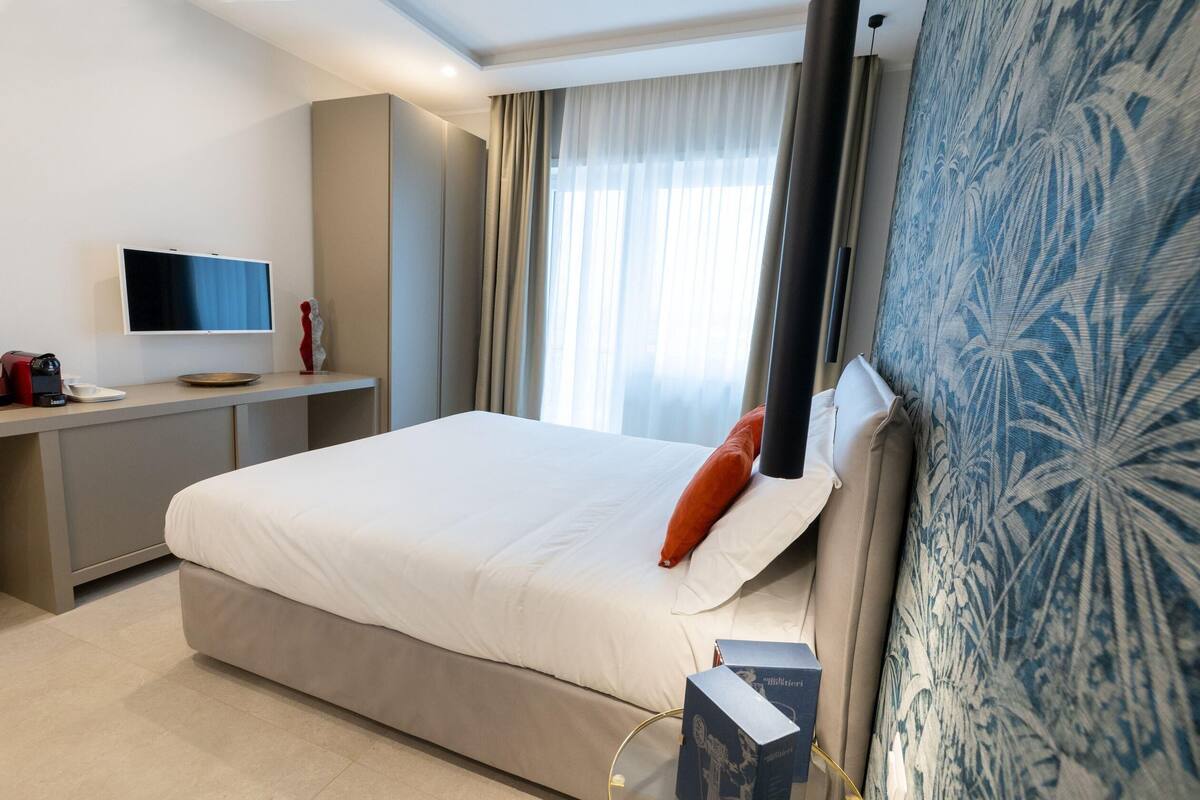 A hotel room with a large bed, a flat-screen TV, a desk with a computer, and a window with curtains.