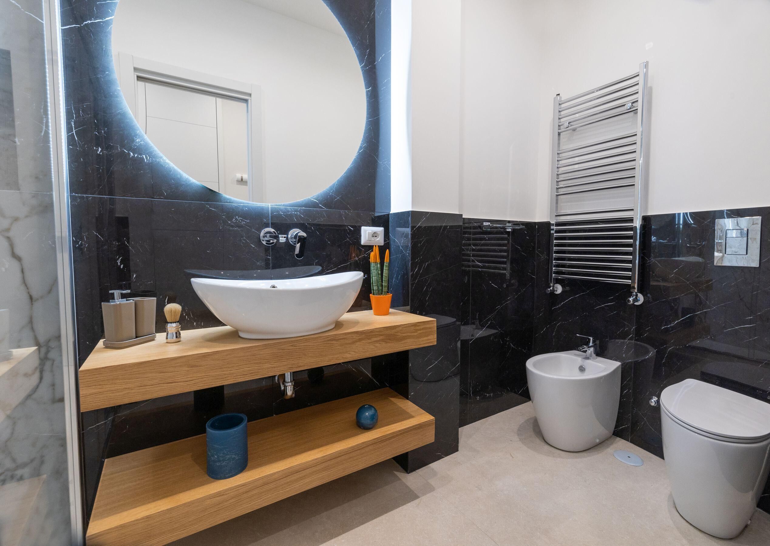 Une salle de bain moderne avec un grand miroir ovale, un lavabo sur colonne blanc, un meuble-lavabo en bois, un porte-serviettes et des toilettes.