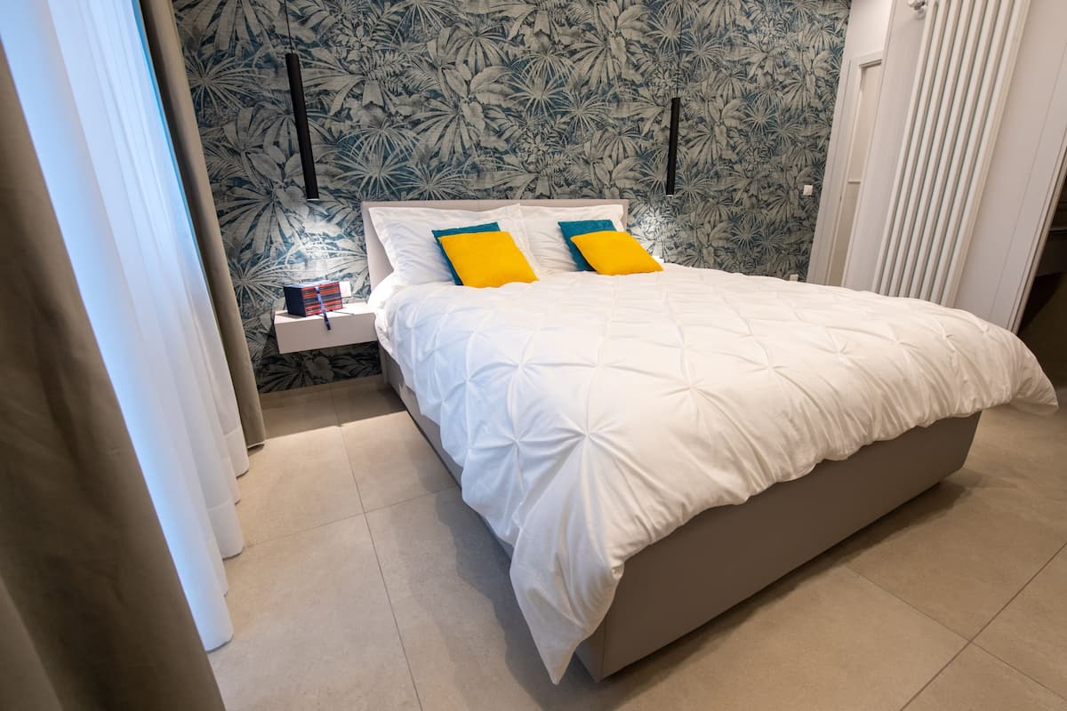 A neatly made bed with white bedding and two yellow pillows, a bedside table with a lamp, and a floral wallpaper pattern on the wall.