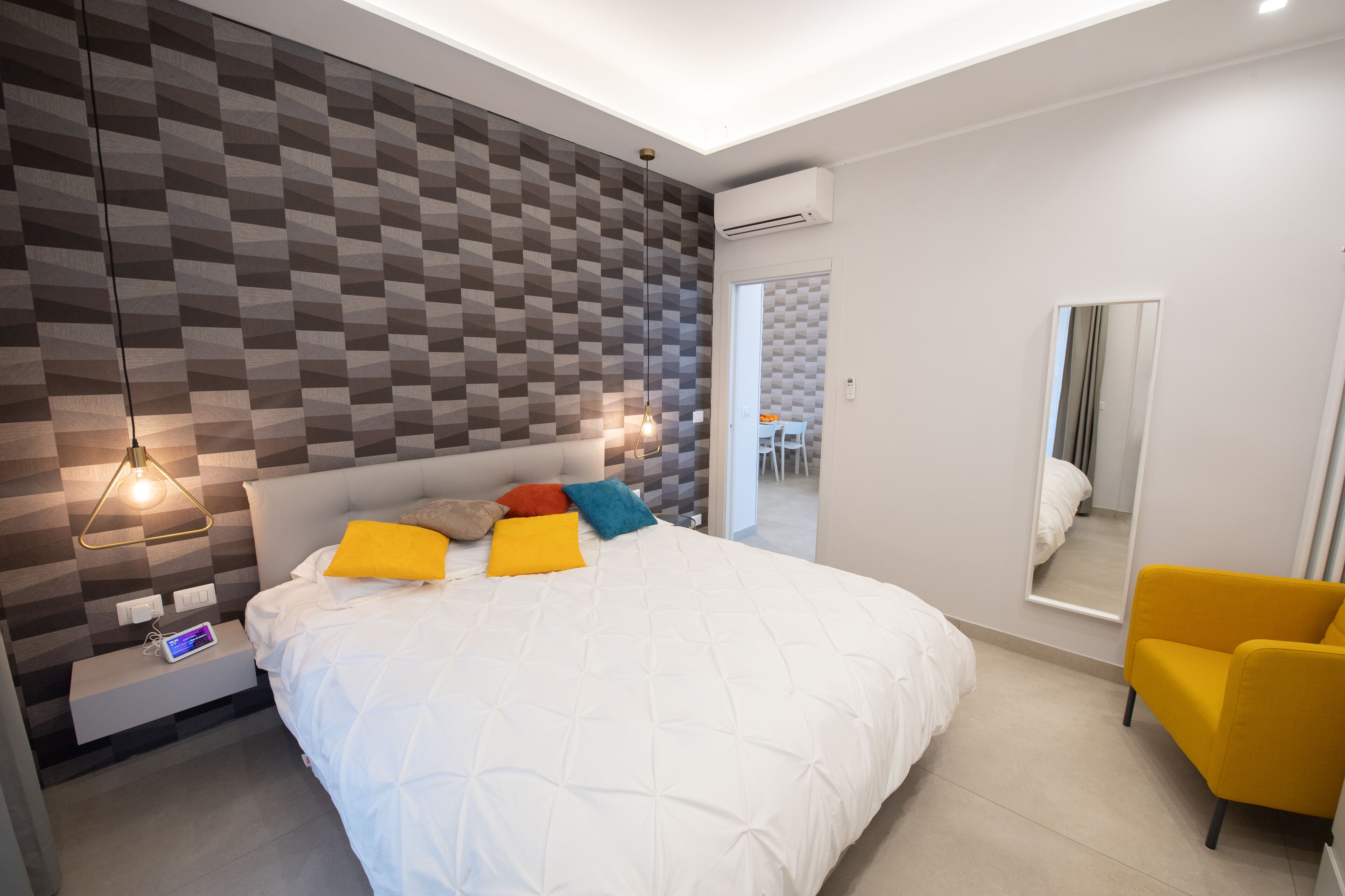 Un dormitorio moderno con pared de estampado geométrico, cama blanca, almohadas coloridas, una mesita de noche con lámpara y un sillón amarillo.