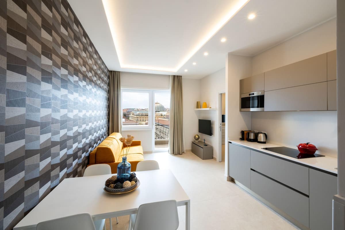 A modern hotel room with a dining area, kitchen, and a view of the cityscape.