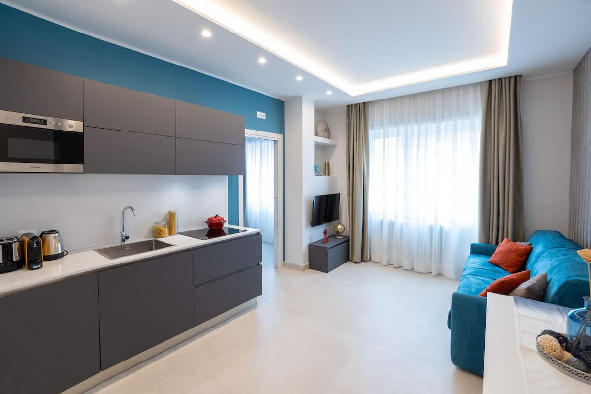 A modern kitchen with grey cabinets, a stainless steel oven, and a sink. There is a blue sofa with cushions in the living area, and a flat-screen TV mounted on the wall.
