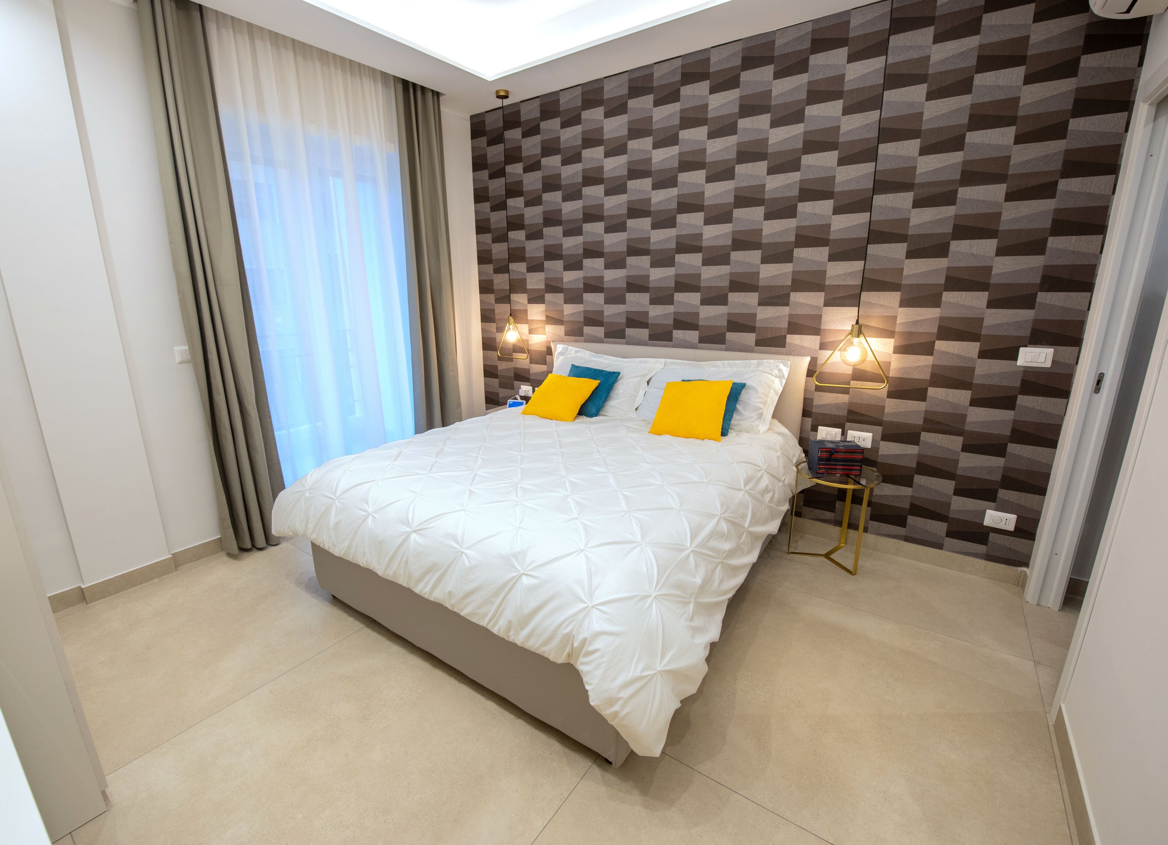 A modern hotel room with a large bed, a geometric-patterned wall, and a bedside table with a lamp.