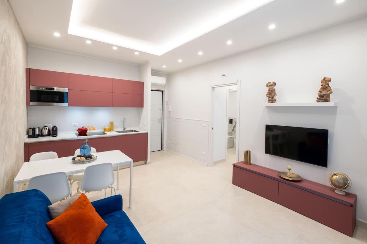 A modern living room with a blue sofa, a flat-screen TV mounted on the wall, and a kitchen area with red cabinets and a microwave.