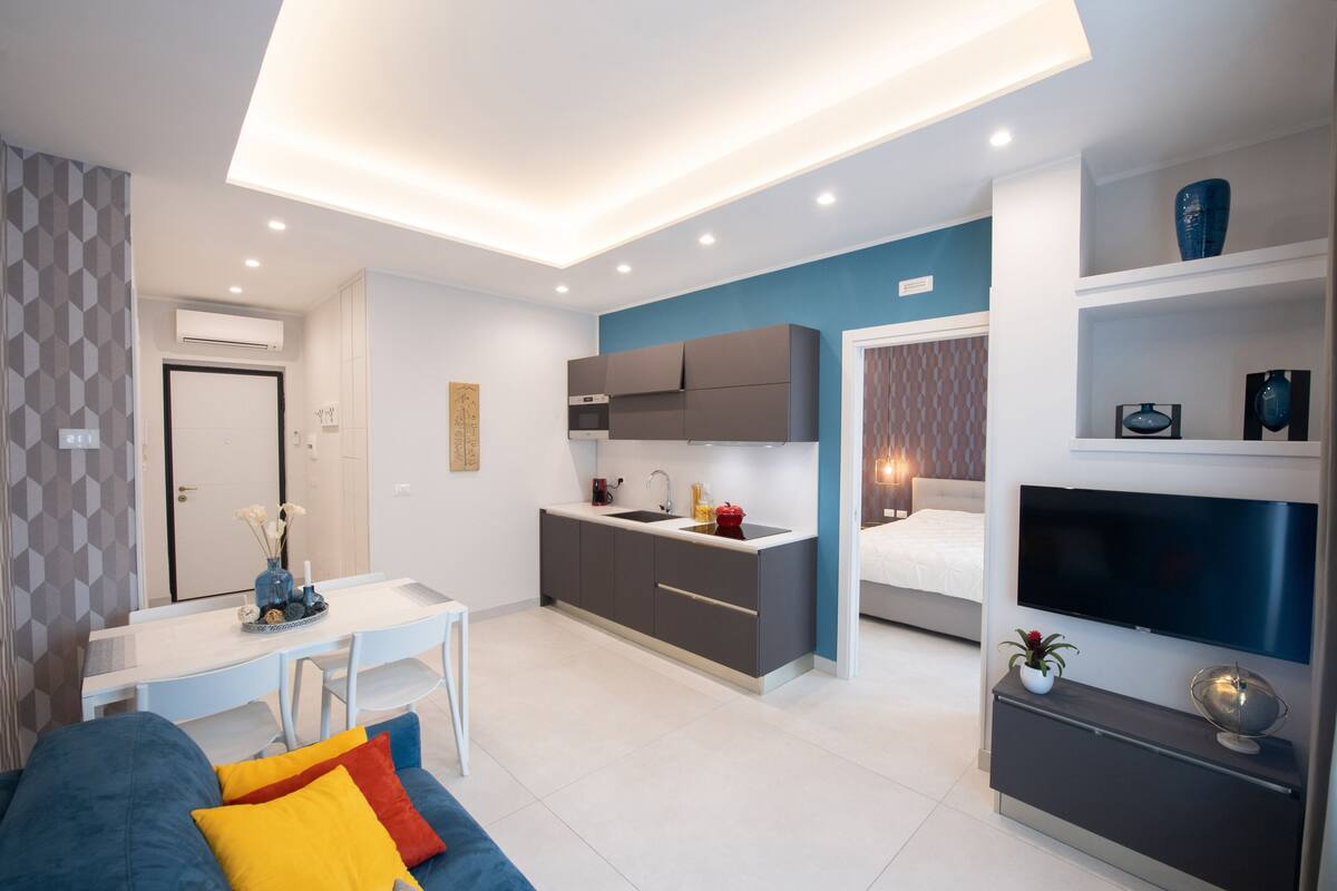 A modern apartment with a kitchen, dining area, and living room. It features a flat-screen TV, a sofa with colorful cushions, and a small side table with decorative items.