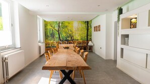 Un coin repas avec une longue table en bois, plusieurs chaises, une fresque représentant une forêt sur le mur et une étagère blanche où sont rangés de la vaisselle.