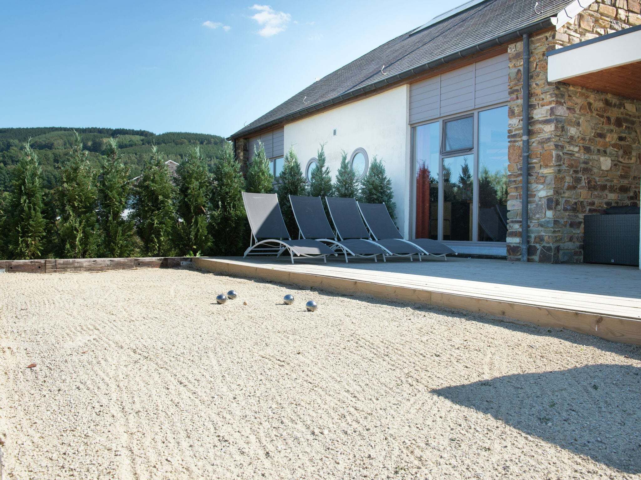 A modern building with a stone facade, a covered patio with lounge chairs, and a view of a forested hillside.