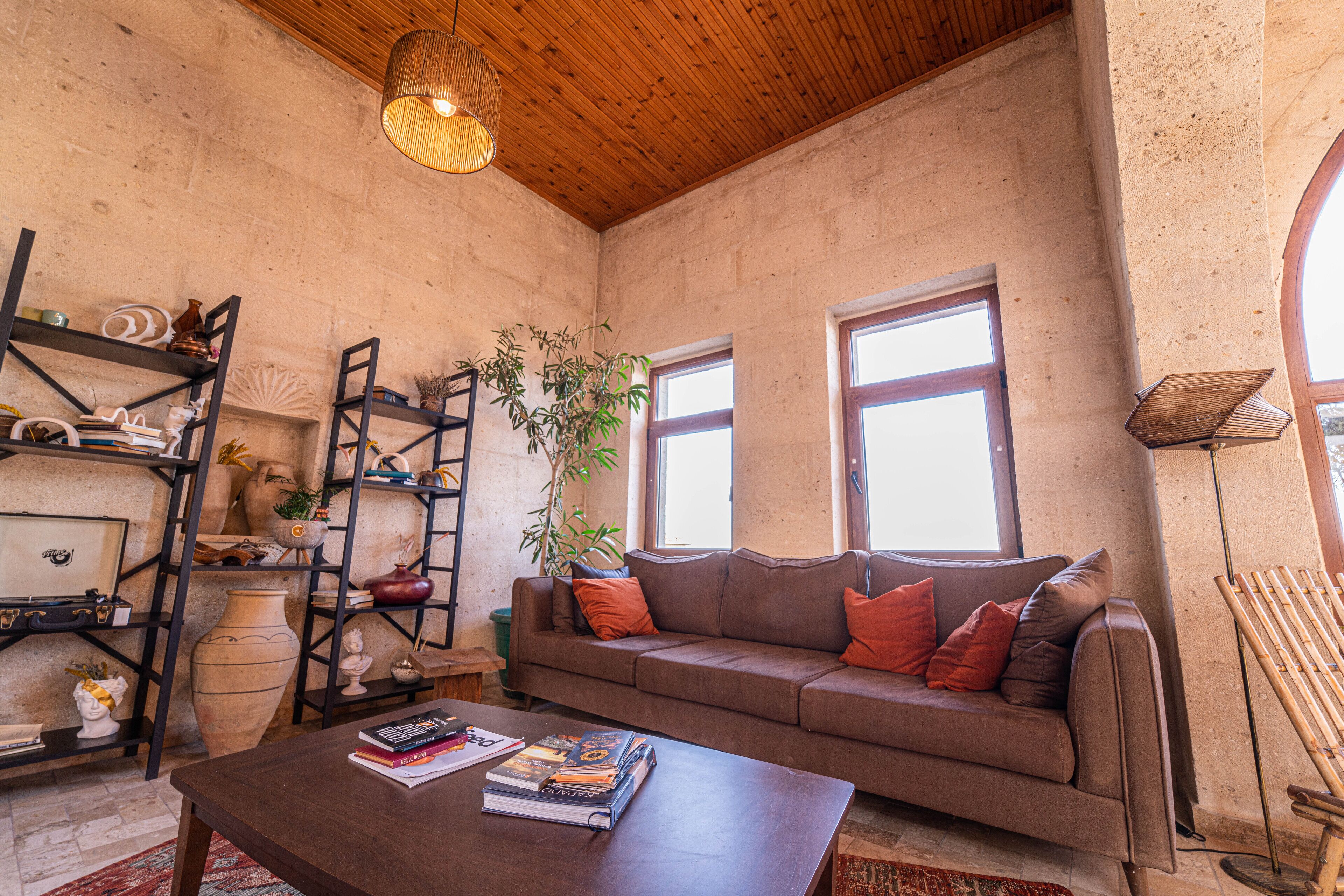 A cozy living room with a wooden ceiling, a brown sofa with orange cushions, a coffee table with books, and a plant.