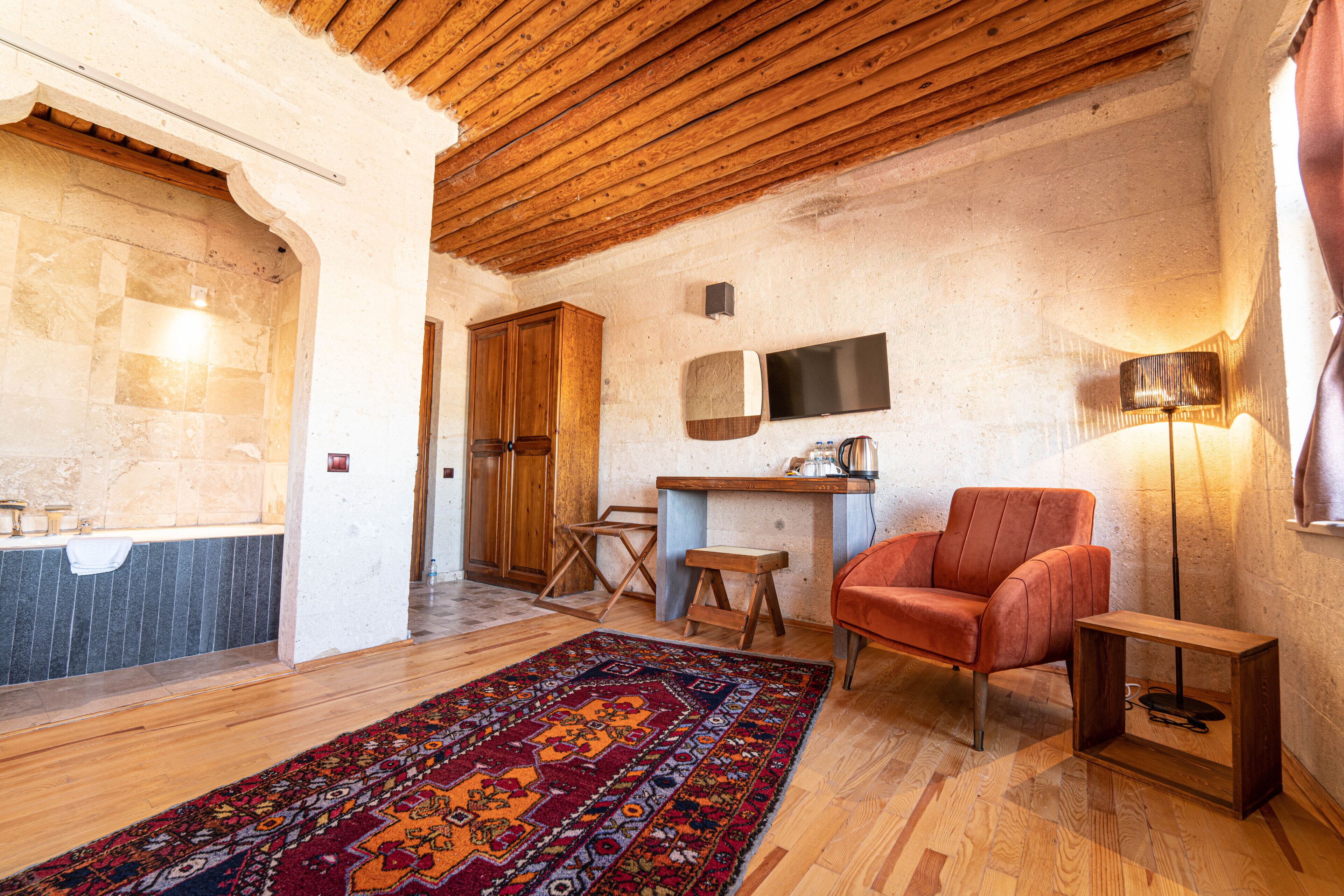 A rustic room with wooden beams, a stone wall, a wooden cabinet, a television, a red chair, a wooden side table, and a patterned rug.