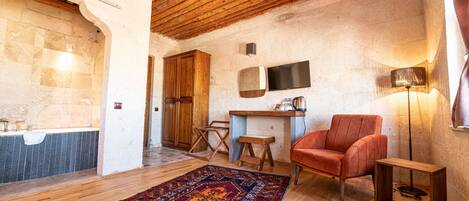 A rustic room with wooden beams, a stone wall, a wooden cabinet, a television, a red chair, a wooden side table, and a patterned rug.