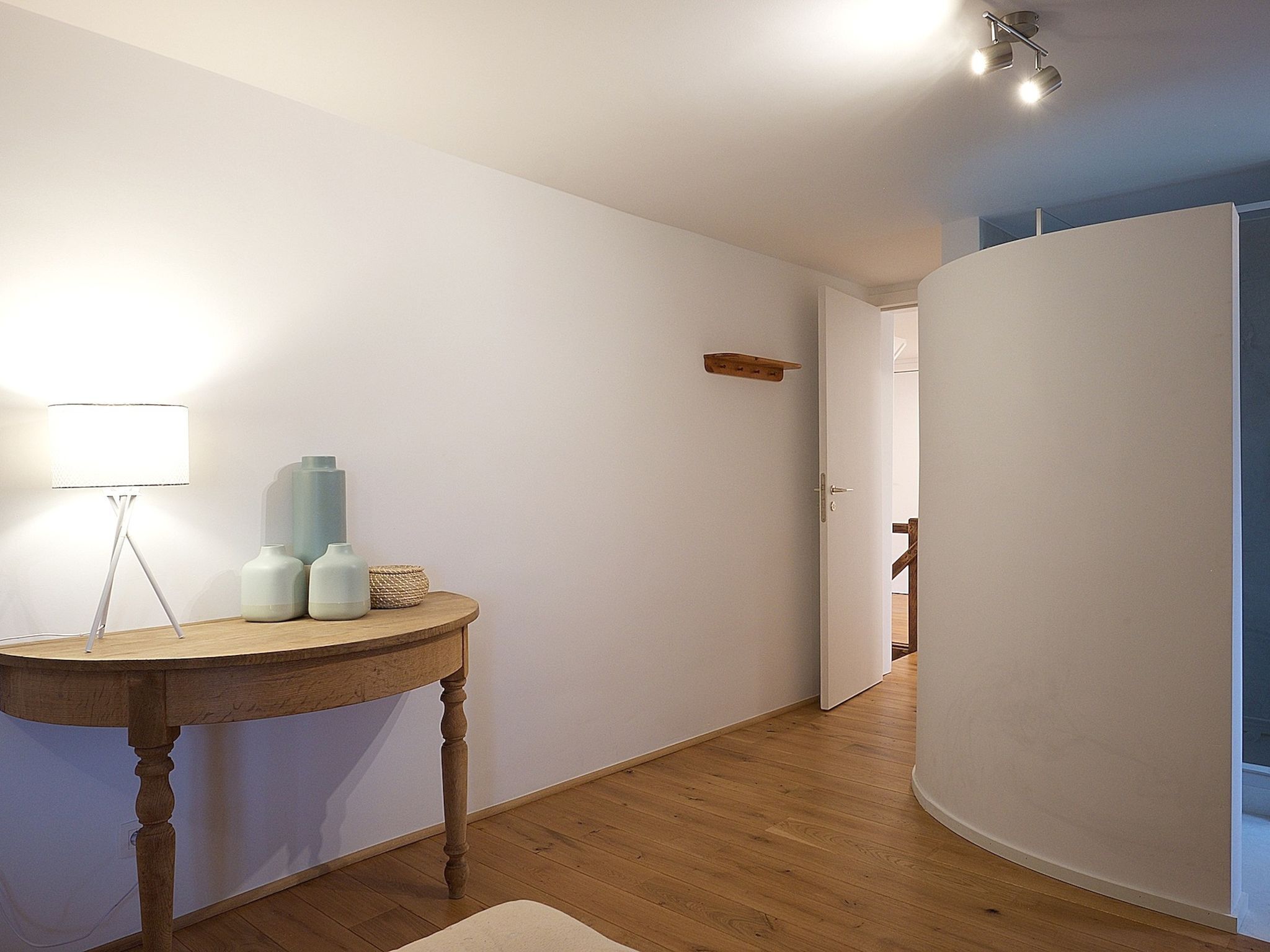 A hallway with a wooden floor, a round table with a lamp, and a door leading to another room.