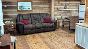 A cozy cabin interior with a wooden floor, a leather sofa, a dining table with wicker chairs, and a refrigerator.