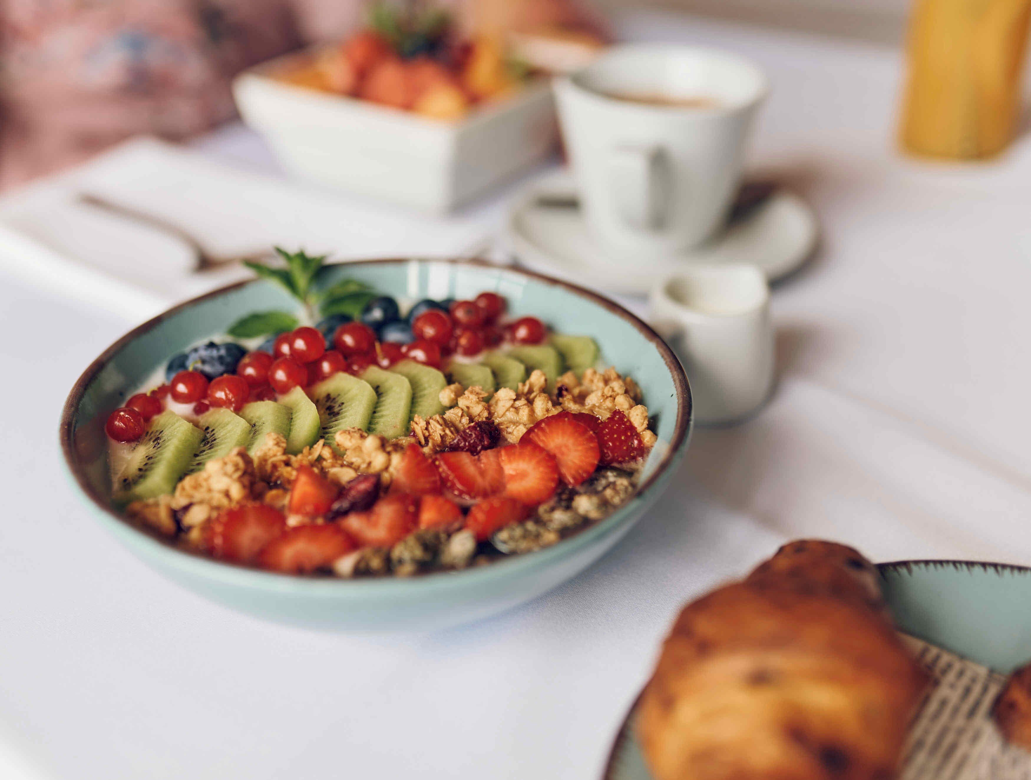 Un desayuno con un bol de fruta y granola, una taza de café y un croissant sobre una mesa.