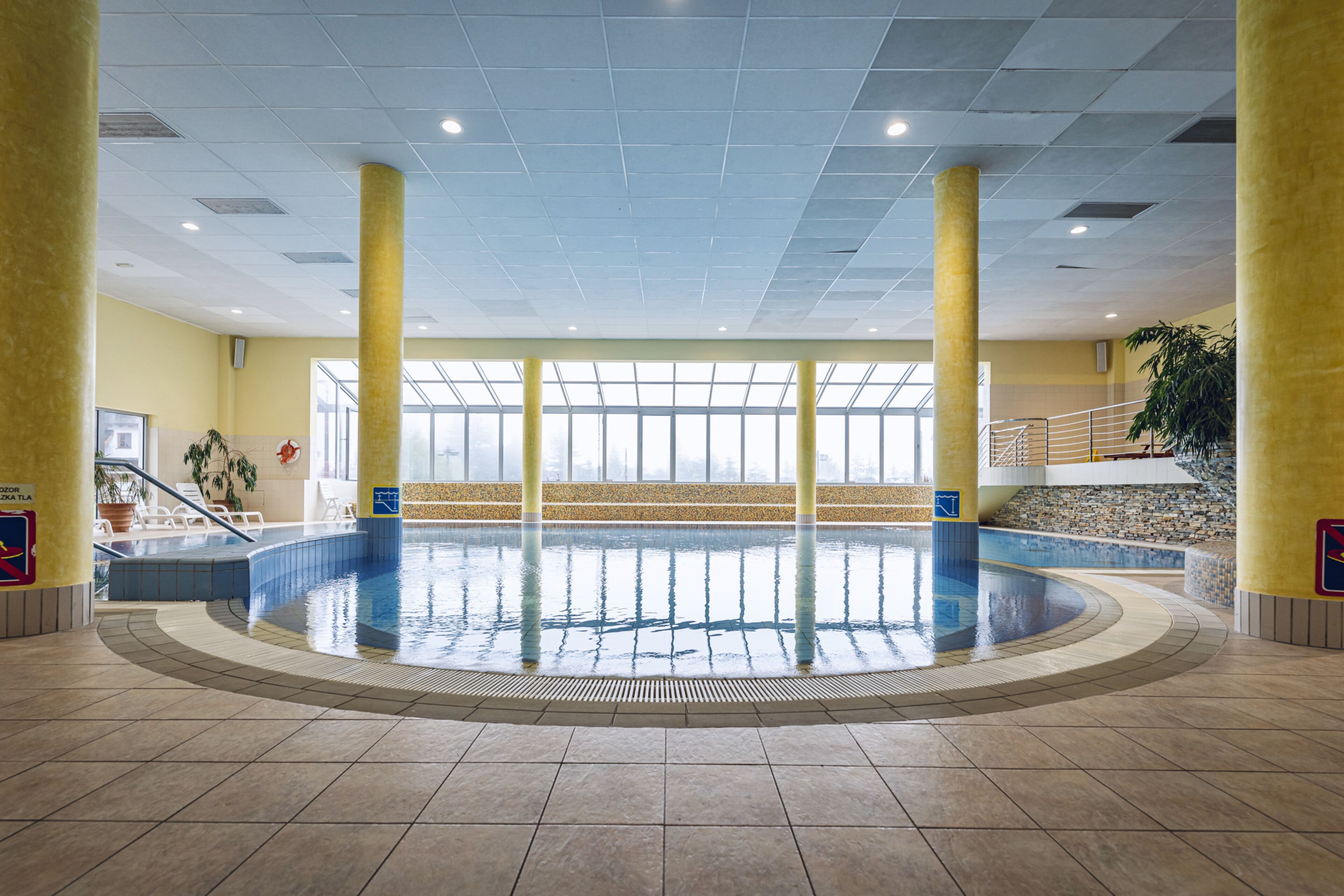 An indoor swimming pool with clear glass walls, yellow pillars, and a tiled floor.