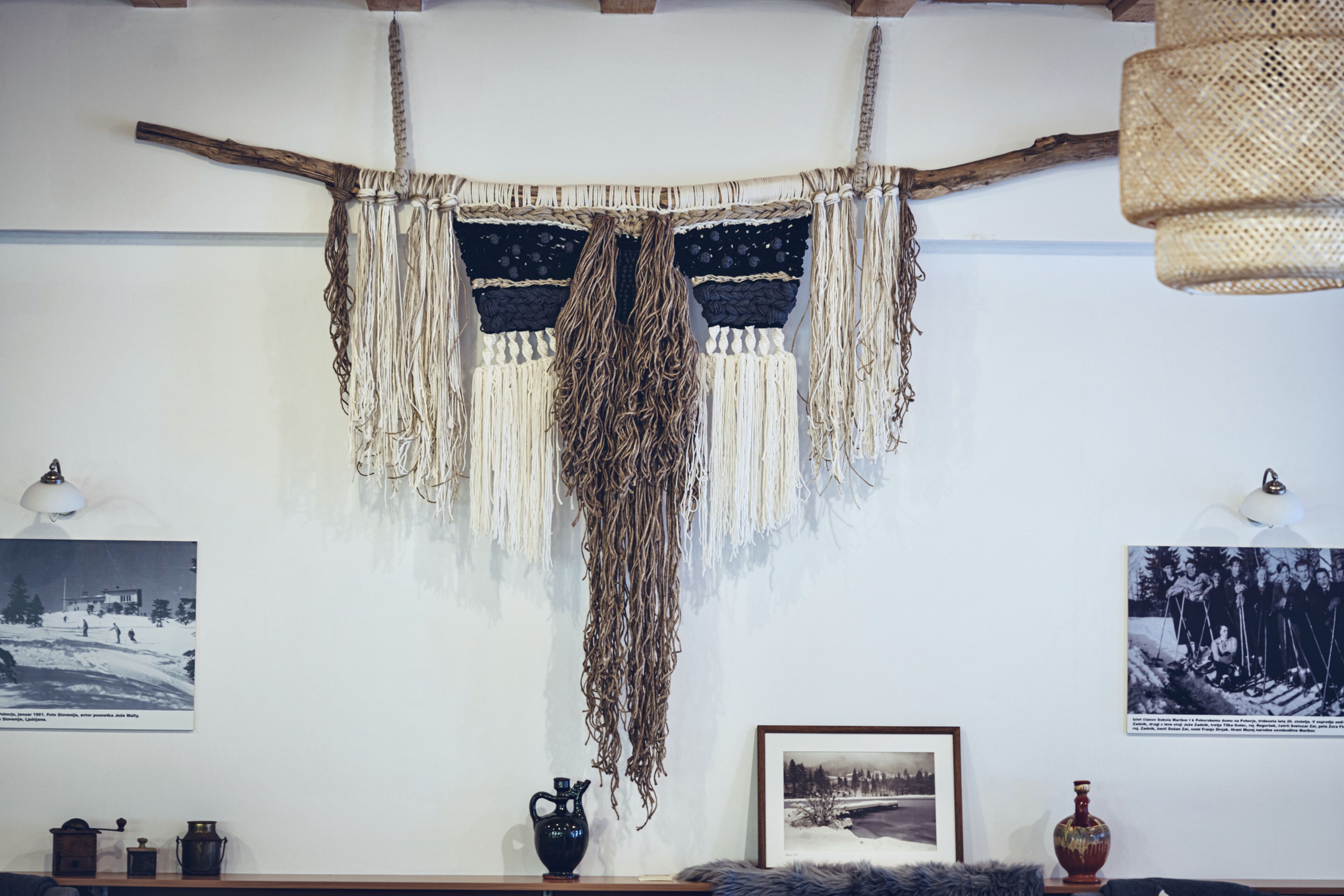 A room with a macrame wall hanging, a wooden shelf with decorative items, and framed photographs on the wall.